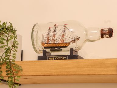 A model ship labeled HMS Victory inside a glass bottle on a wooden shelf with a plant nearby at Lakeside in Falmouth