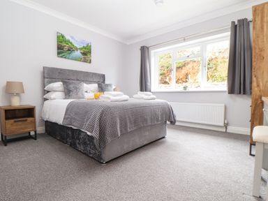 A bedroom with a grey bed a bedside table with a lamp a window with curtains and a picture above the bed at Lakeside in Falmouth