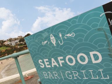 A seafood bar and grill sign on a railing by the beach with houses on a hill in the background at Lakeside in Falmouth