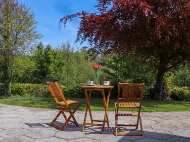 An outdoor patio with a wooden table and two wooden chairs with red flowers in a vase on the table at Lakeside in Falmouth
