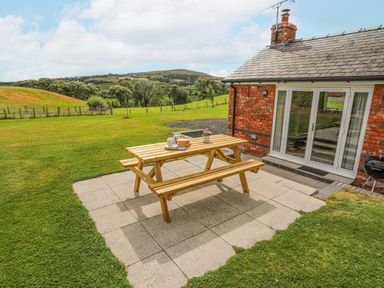 A wooden picnic table with dishes on a paved patio next to a brick building in a grassy field at The Piggery in LLanfair Talhaiarn near Abergele