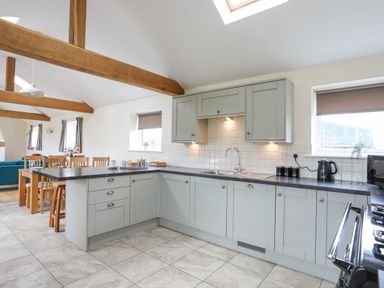 A kitchen with grey cabinets a black countertop a stove and dining table with chairs and wooden beams on ceiling at The Piggery in LLanfair Talhaiarn near Abergele