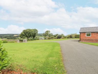 A paved driveway next to a red brick building with a grassy lawn and trees in the background at The Piggery in LLanfair Talhaiarn near Abergele