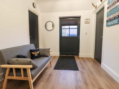 An entryway with a wooden framed gray sofa with cushions a black door a black mat on wood flooring and two black doors on the sides at Bee Cottage in Ottinge near Lyminge