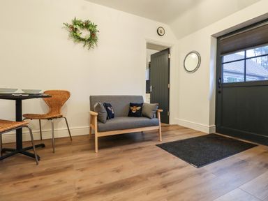 A room with a small grey sofa with patterned cushions a black door a round wall mirror a dining table with two wicker chairs and wooden flooring at Bee Cottage in Ottinge near Lyminge