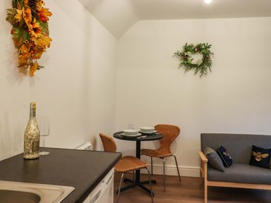 A small dining area with a round table set for two wicker chairs a gray sofa with decorative cushions and a kitchen counter with a bottle and glass at Bee Cottage Ottinge near Lyminge