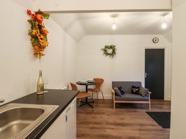 A room with a sink and countertop with a bottle and glass a round table with two chairs a gray loveseat with pillows a black door and wall decorations at Bee Cottage in Ottinge near Lyminge