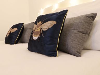 Two decorative pillows with bee embroidery and two plain grey pillows on a bed at Bee Cottage in Ottinge near Lyminge