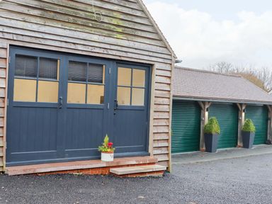 A wooden building with blue double doors and a potted plant next to a row of green garage doors with topiary plants in tall black pots at Bee Cottage Ottinge near Lyminge