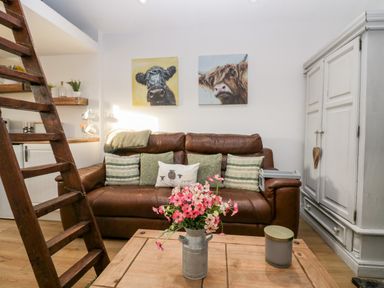 A living room with a brown leather sofa cushions paintings of cows a wooden ladder a wooden coffee table with flowers and a candle and a white cabinet at High Wreah Bolt Hole in Cleator Moor