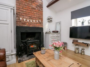 A living room with a brick fireplace a wooden coffee table with flowers a mounted TV and a white chest of drawers at High Wreah Bolt Hole in Cleator Moor