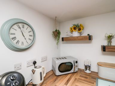A kitchen corner with a wall clock, kettle, microwave, hanging plant, sunflowers on a shelf, cutlery holder, and bread bin at High Wreah Bolt Hole in Cleator Moor