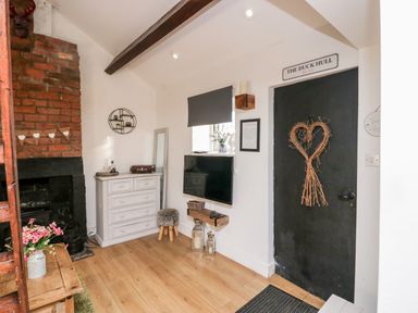 A small sitting area with a black door decorated with a heart-shaped wreath wooden floor a white chest of drawers a wall-mounted TV and a brick fireplace at High Wreah Bolt Hole in Cleator Moor