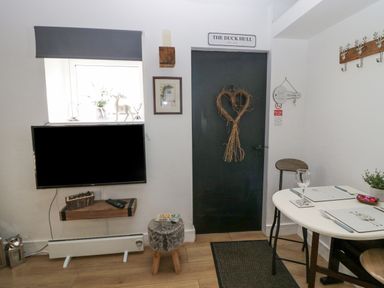 A dining area with a table set for two near a black door with a heart-shaped wreath and a wall-mounted television at High Wreah Bolt Hole in Cleator Moor