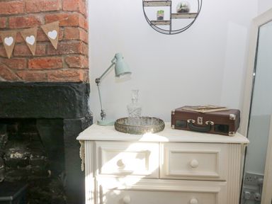 A white dresser with a lamp a tray with glassware and a brown suitcase next to a brick fireplace at High Wreah Bolt Hole in Cleator Moor