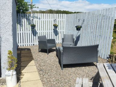 An outdoor seating area with three grey chairs and a glass table on gravel next to a white wooden fence with hanging flower pots at High Wreah Bolt Hole in Cleator Moor