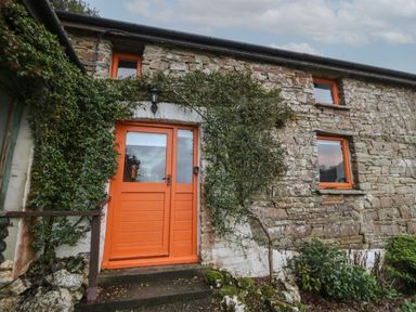 An exterior view of a stone building with orange framed windows and an orange door surrounded by green plants at Ty Ceffylau in Llanllwni near Llanybydder