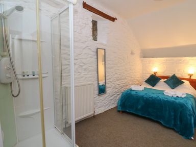 A bedroom with a double bed with blue blanket and pillows a shower cubicle and a mirror on a white stone wall at Y Beudy in Cwmiar near Llanybydder