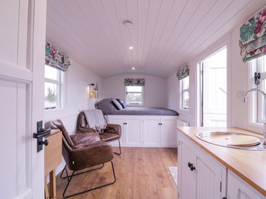 A small room with a bed elevated over cabinets two leather chairs a wooden floor a kitchenette with a sink and windows with floral curtains at Bumbles Retreat in Whepstead near Bury St Edmunds