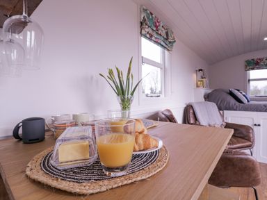 A dining table with croissants, butter, orange juice and a vase with flowers in a room with two windows at Bumbles Retreat in Whepstead near Bury St Edmunds