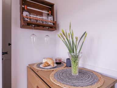 A sideboard with a vase of flowers and croissants on plates under hanging wine glasses at Bumbles Retreat in Whepstead near Bury St Edmunds