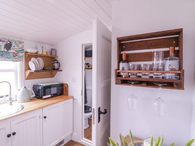 A kitchen countertop with a microwave kettle and shelving holding dishes and mugs with a wooden rack holding gin bottles and glasses at Bumbles Retreat in Whepstead near Bury St Edmunds
