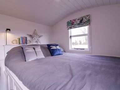 A bedroom with a large bed with gray bedding two dragonfly cushions a window with a floral blind and a star decoration on the wall at Bumble's Retreat in Whepstead near Bury St Edmunds