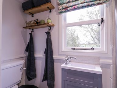 A bathroom with a toilet a sink two hanging towels wooden shelves and a window with patterned blind at Bumbles Retreat in Whepstead near Bury St Edmunds