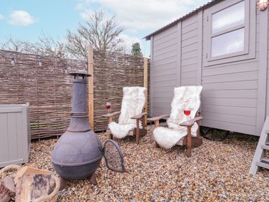 An outdoor seating area with two wooden chairs covered with fur throws a black chiminea and a basket of firewood at Bumble's Retreat in Whepstead near Bury St Edmunds