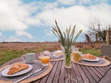 Breakfast setting with croissants mugs and glasses of orange juice on a wooden table outdoors with a vase of flowers at Bumble's Retreat in Whepstead near Bury St Edmunds