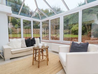A conservatory with glass ceiling and windows containing a sofa and armchair with cushions and a small wooden table with cups and teapot at Sunningdale Rise in Preston