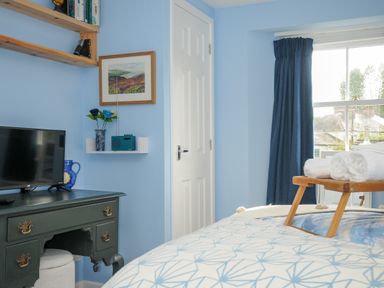 A bedroom with a bed, a black dresser with a television, a small white shelf with a blue radio and a vase with flowers, and a window with dark curtains at 15 The Moors in Lostwithiel