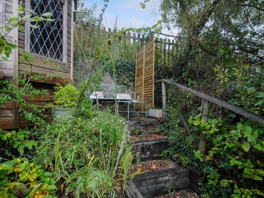 An outdoor garden seating area with wooden steps leading up to a metal bench and table surrounded by plants and bushes at 15 The Moors in Lostwithiel