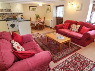 A living room with red sofas and cushions a wooden coffee table and rugs next to a dining table and kitchen area at Swallow Cottage in Mawgan