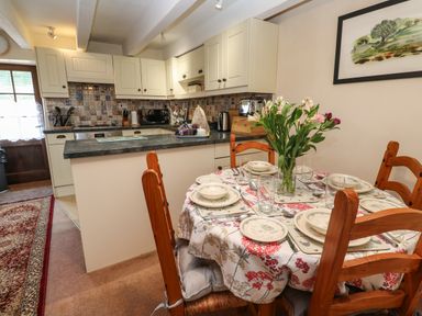 A dining area with a round table set for six with plates and glasses and a vase of flowers next to a kitchen with cream cabinets and tiled backsplash at Swallow Cottage in Mawgan