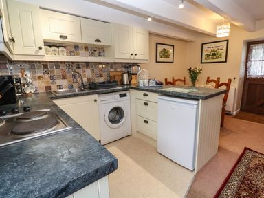 A kitchen with white cabinets a washing machine a small refrigerator a dining table with chairs and flowers at Swallow Cottage in Mawgan