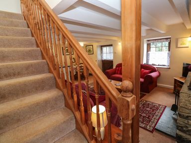 A carpeted staircase with wooden railing leading to a living area with red sofas a coffee table a small rug a wood stove and a window with curtains at Swallow Cottage in Mawgan