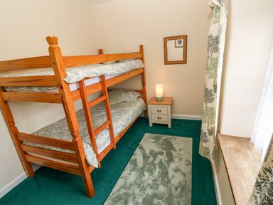 A bedroom with wooden bunk beds a nightstand with a lamp a mirror on the wall a window with curtains and a carpet on the floor at Swallow Cottage in Mawgan