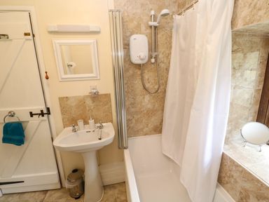 A bathroom with a pedestal sink a bathtub with shower and curtain a mirror a towel on door and a small round mirror on a tiled ledge at Swallow Cottage in Mawgan