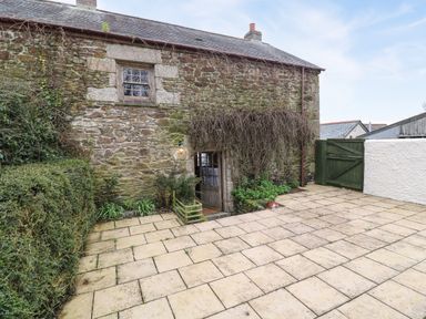 A stone cottage with a small wooden door covered by branches and a paved patio with a green gate at Swallow Cottage in Mawgan
