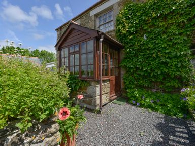Stone house with wooden porch surrounded by plants and flowers at Swallow Cottage in Mawgan