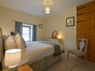 A bedroom with a bed pillows curtains a wooden chair a chest of drawers with mirror a lamp and wooden wardrobes at Swallow Cottage in Mawgan