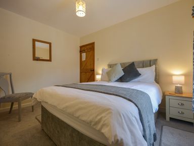 A bedroom with a bed with pillows and blanket a wooden door a mirror a bedside table with lamp and a chair at Swallow Cottage in Mawgan