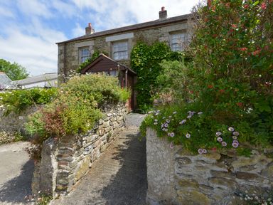 A stone cottage with a wooden entrance surrounded by flower beds and climbing plants at Swallow Cottage in Mawgan