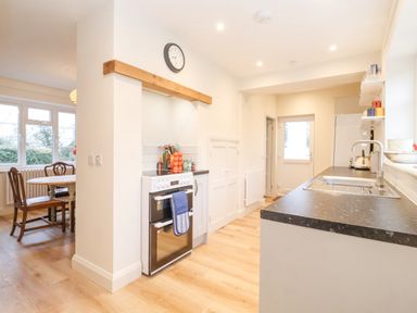 A kitchen with a stove, countertop with sink, shelves with glasses, and a dining area with wooden chairs and table at 3 Chantry Cottages Campsea Ashe near Woodbridge