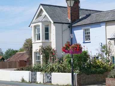 A street view of a two-story white cottage with a garden and hanging flowers at The Corner Cottage in Aldeburgh