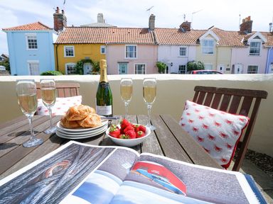 An outdoor wooden table with croissants on plates strawberries a bottle and glasses of champagne and an open book with colorful houses in the background at The Corner Cottage in Aldeburgh