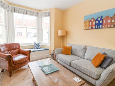 A living room with a gray sofa and orange cushions a leather armchair and a wooden coffee table at The Corner Cottage in Aldeburgh