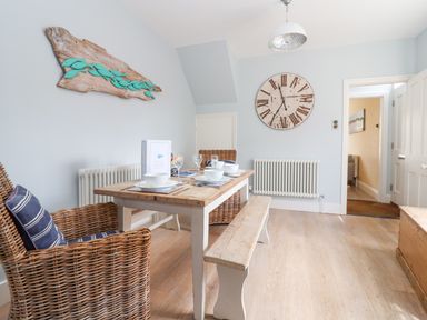 A dining area with a wooden table set for four with wicker chairs and a bench at The Corner Cottage in Aldeburgh