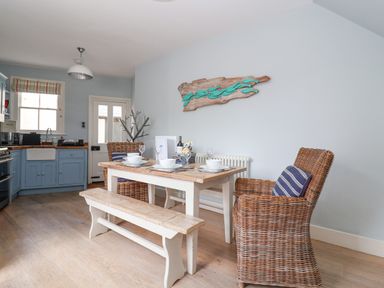 A dining area with wooden table, bench, wicker chairs, and kitchen cabinets at The Corner Cottage in Aldeburgh